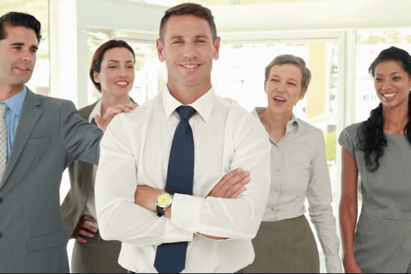 five business professionals with one man in front looking pleased and the others seeming proud of him